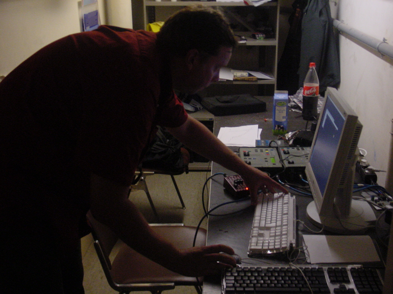Tom leans over a desk with a computer, keyboard and technical equipment in a dark room