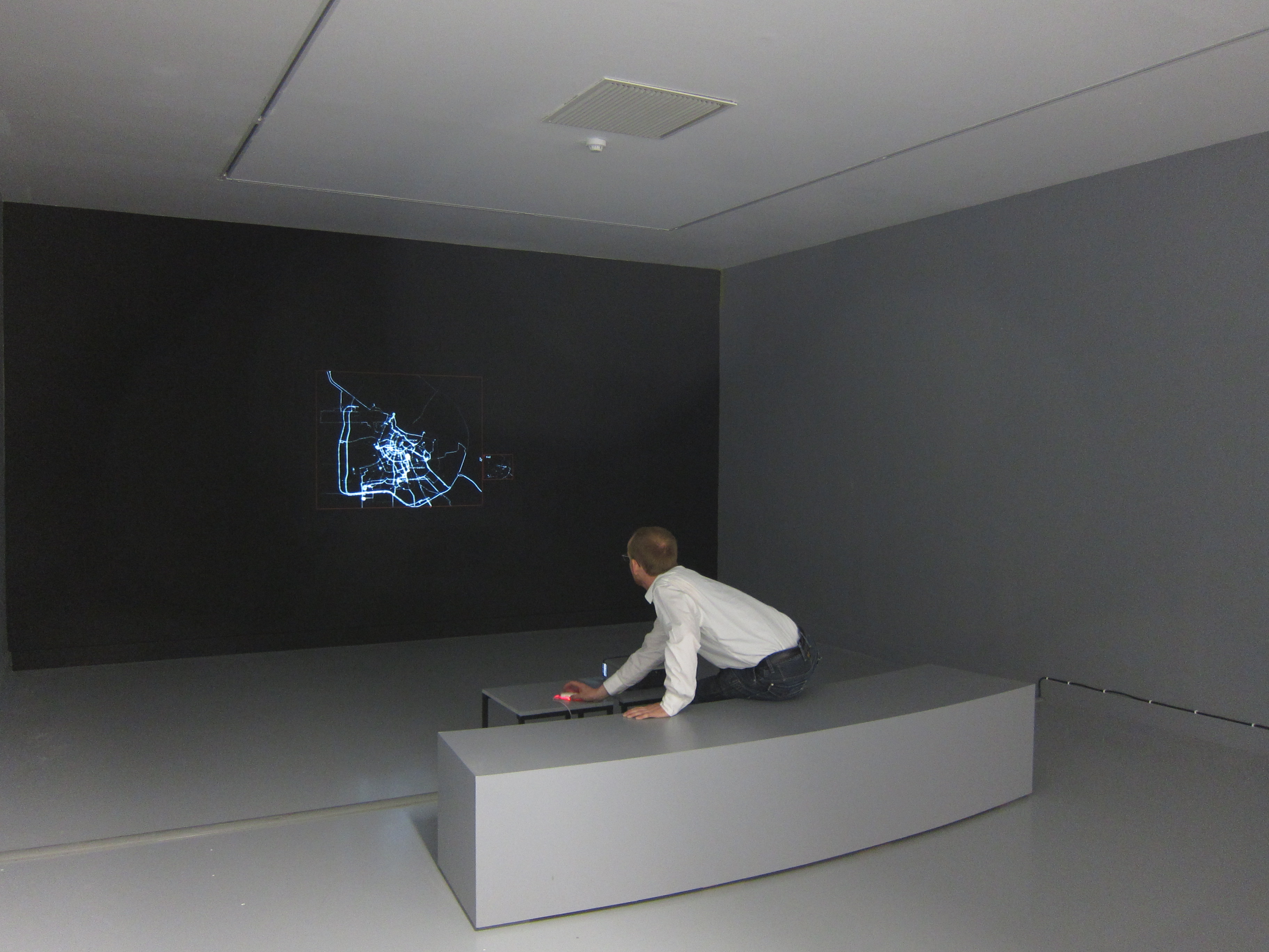 Grey museum space with a projection of blue GPS routes on a dark wall, a person leans forward to operate a laptop