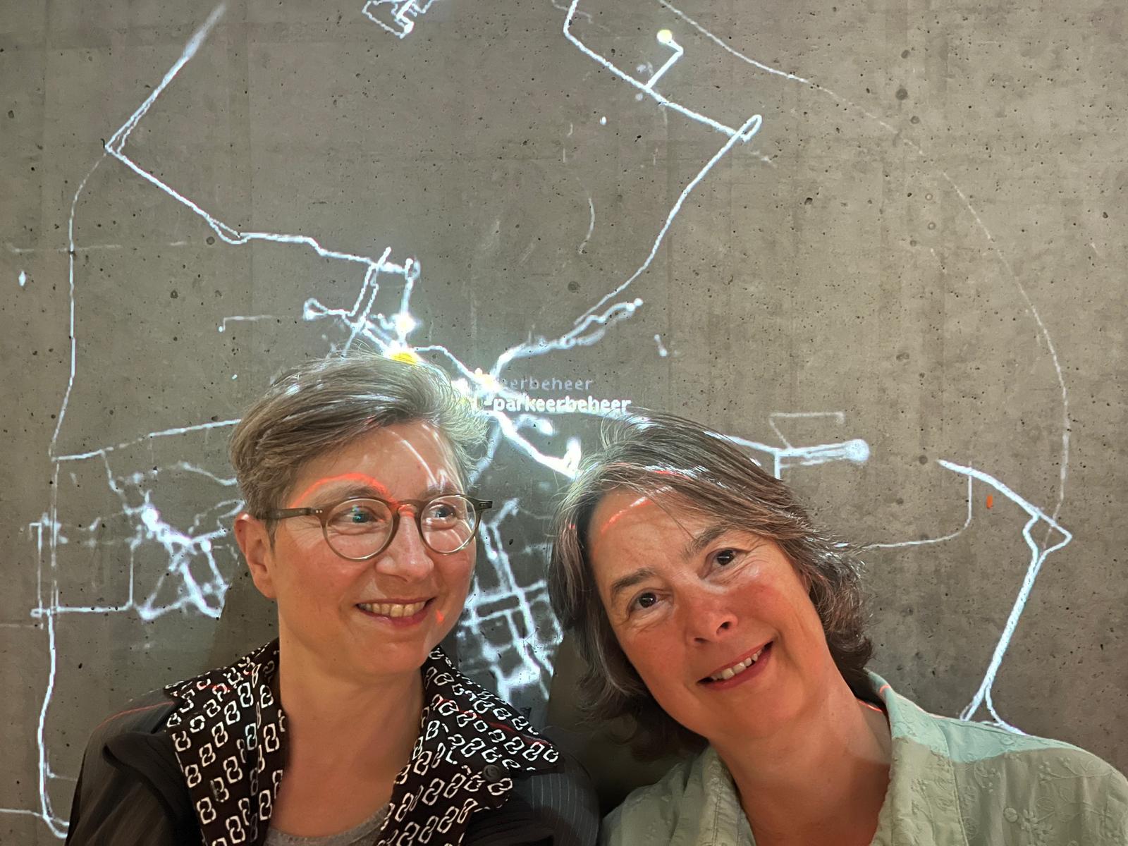 Two women pose smiling in front of a concrete wall onto which a GPS route map is projected