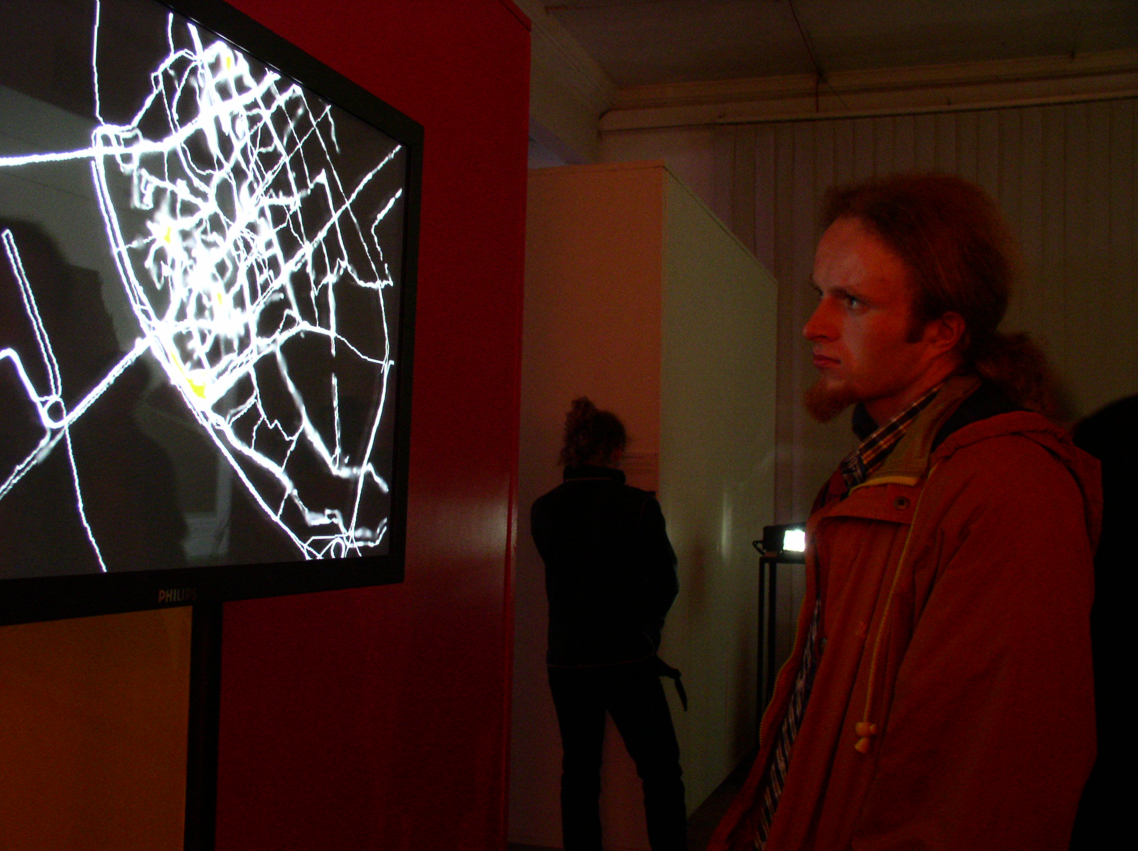 Visitor looks at a screen showing white GPS routes of Riga, in a red-lit exhibition space