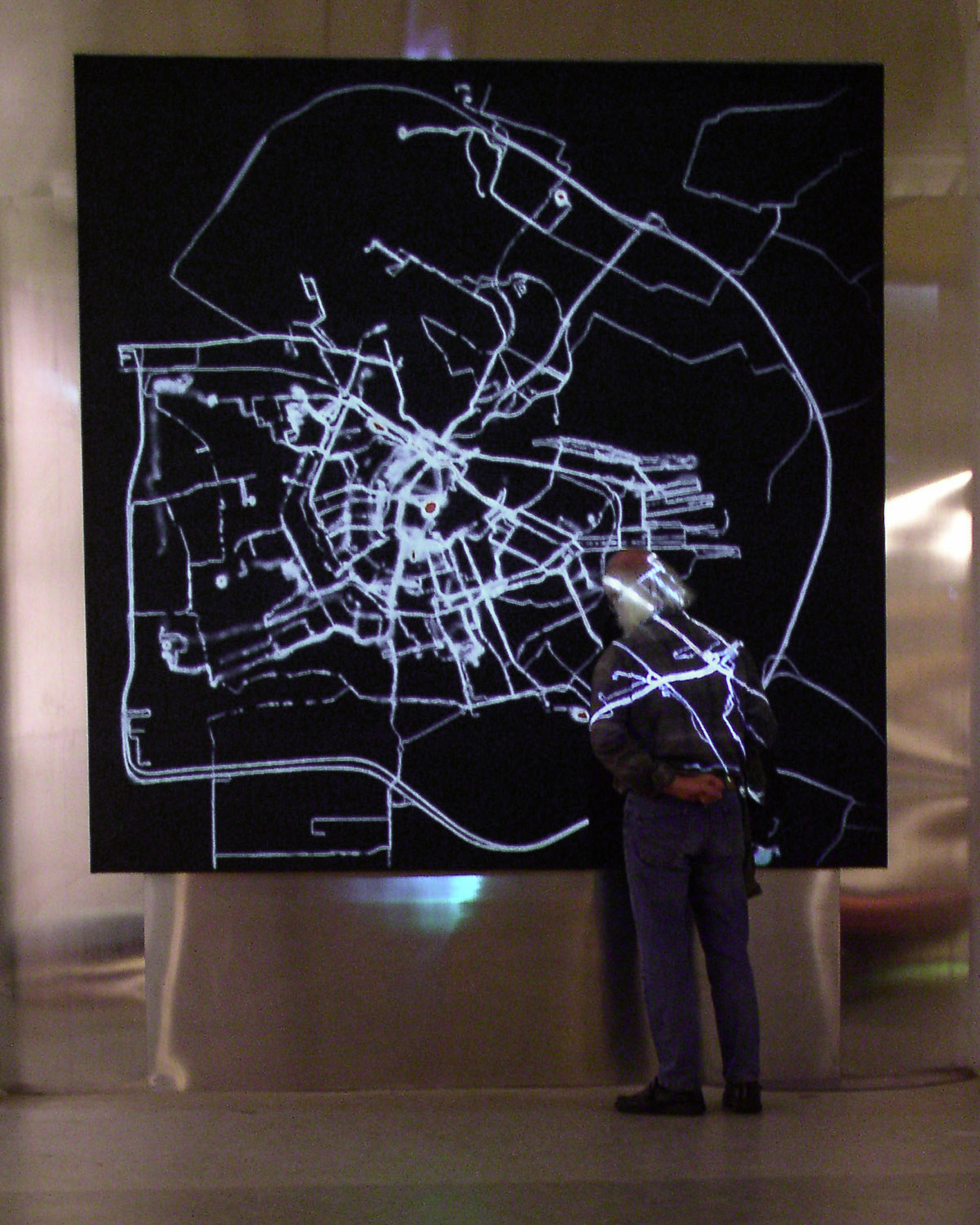 A visitor stands in a dark room in front of a large screen with a projection of white GPS routes revealing the shape of Amsterdam